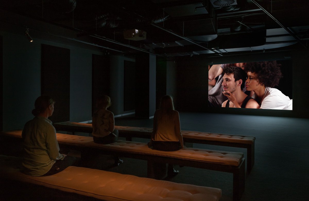 A darkened gallery with three empty bench seats. A moving image work of a black woman with her arms crossed and a white man beside her holding his chest. They appear mid-dance.