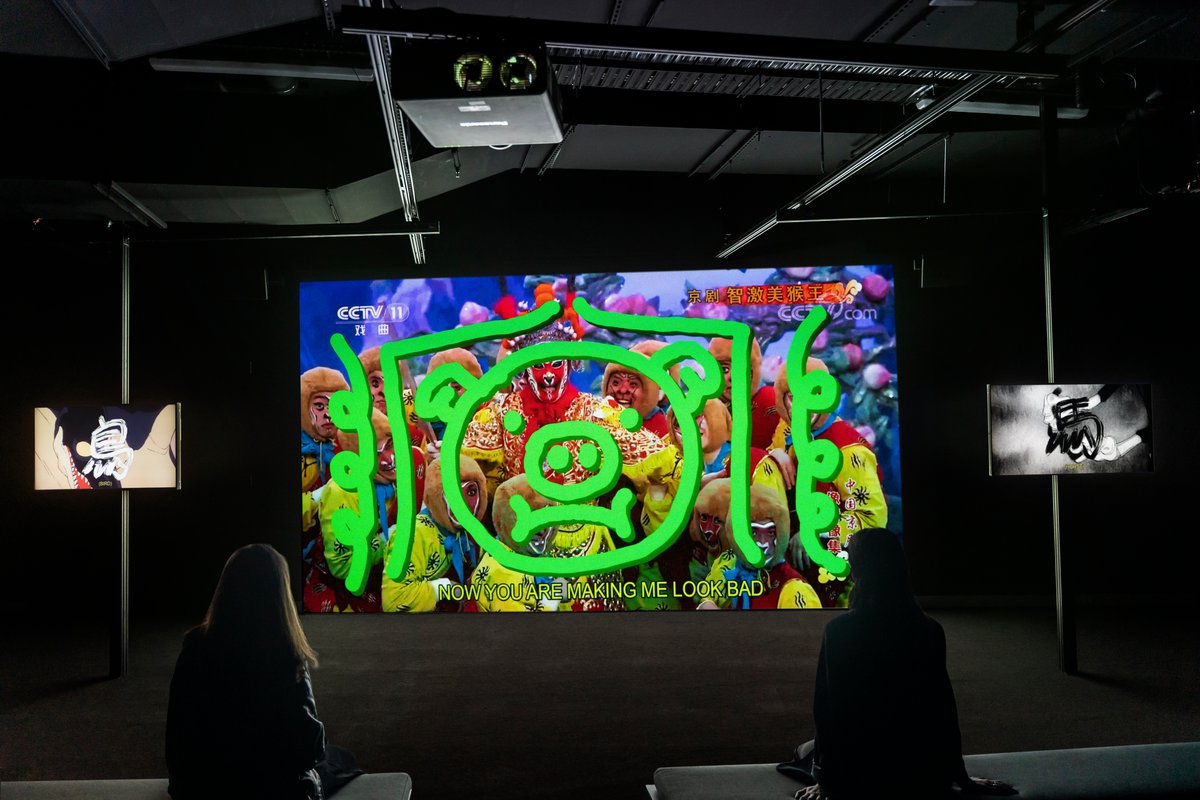 An image of a three-screen work featuring two people watching it, the main screen is colourful and has a animated pig face over smiling people and subtitles reading now you are making me look bad