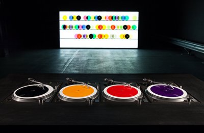 An image of a turntable set up with colourful record records in the background. The four turntables are black, yellow and purple.