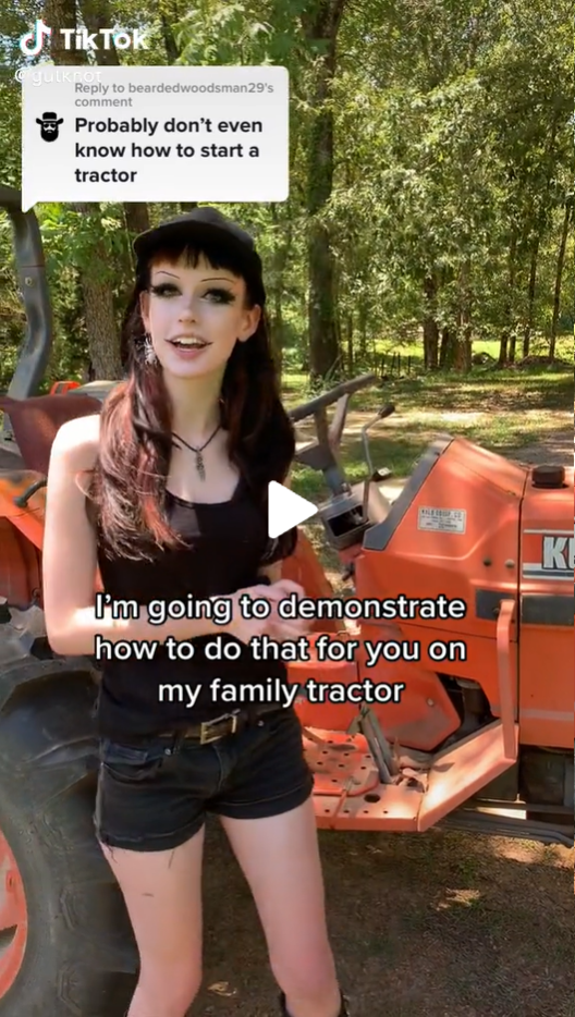 A young woman with dark hair and clothing standing in a rural setting in front of a green tractor