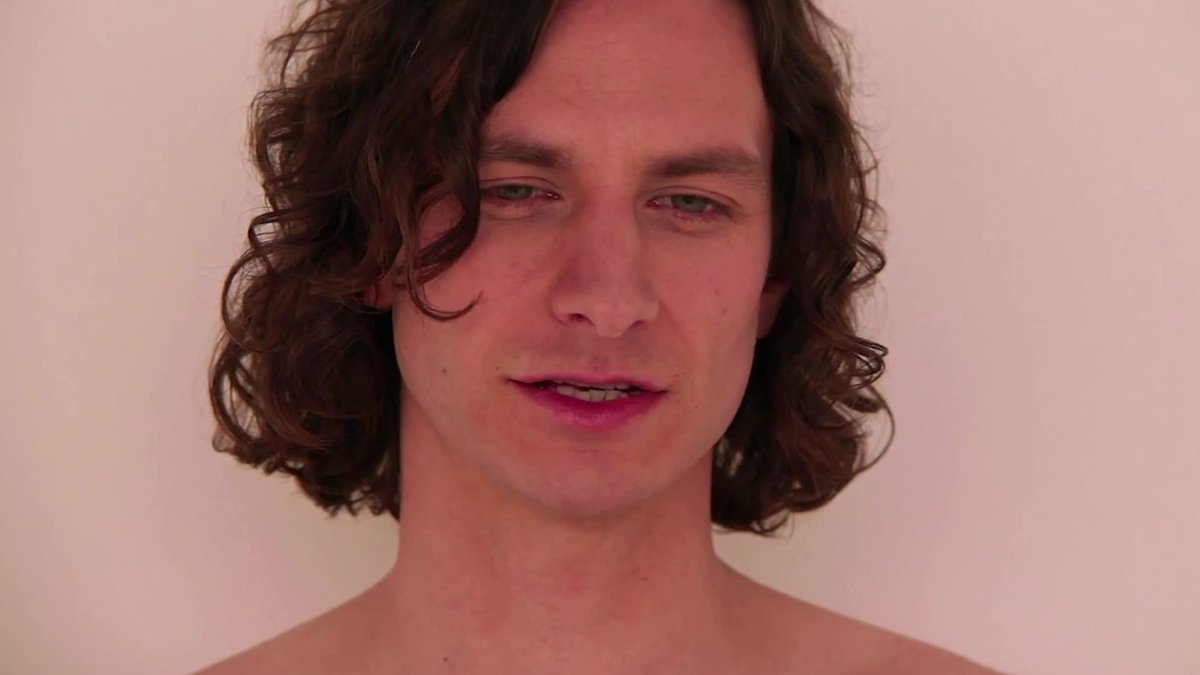 A close-up of Gotye, who has slightly pink lips and chestnut curly shoulder length hair, he's squinting