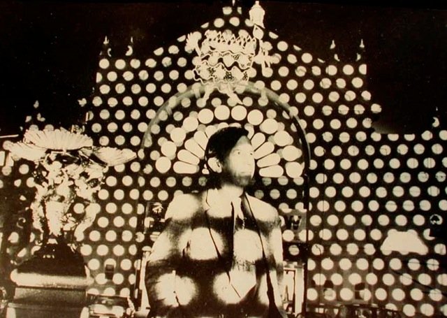 A sepia image of a man standing outside an ornate, grand exhibition building covered in polka dots projected from light