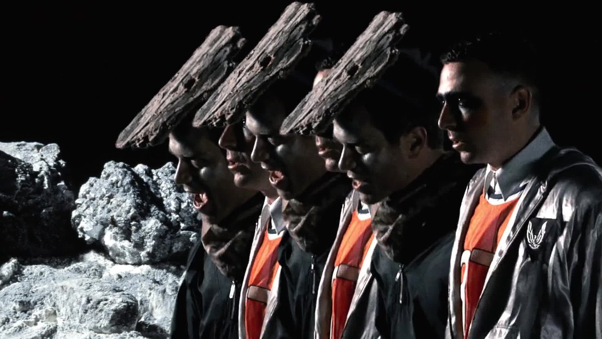 An image of four band members in space age costumes, three of them with rocks on their heads, on a lunar landscape