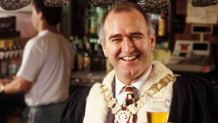 A white man in ceremonial, mayoral robes in a pub drinking a glass of beer.