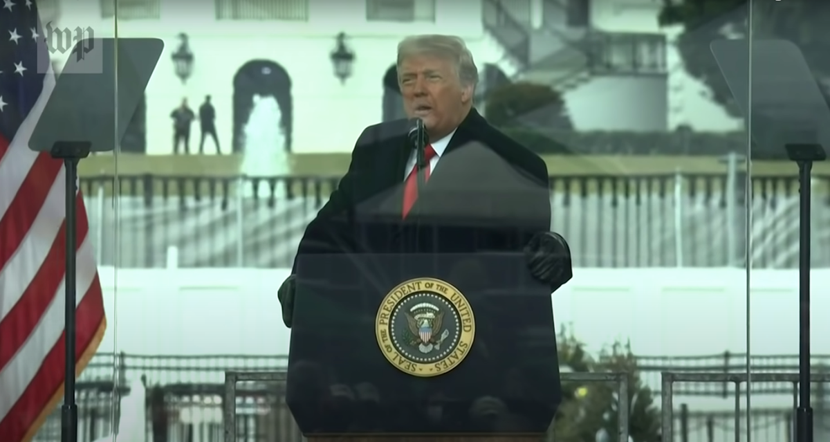 A still frame from a video fo Donald Trump standing before the capitol building on a stage, speaking at a rally, wearing a black coat and gloves, his orange face gleaming arrogantly