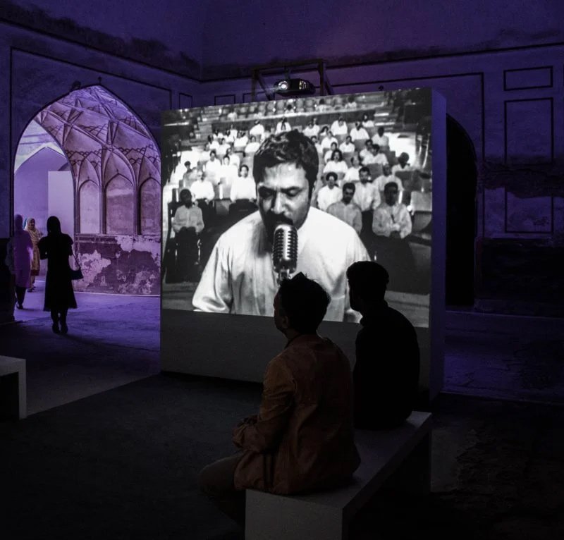 An installation view of a screen-based artwork that features a black and white image of a bearded man, with people seated in a purple room watching it