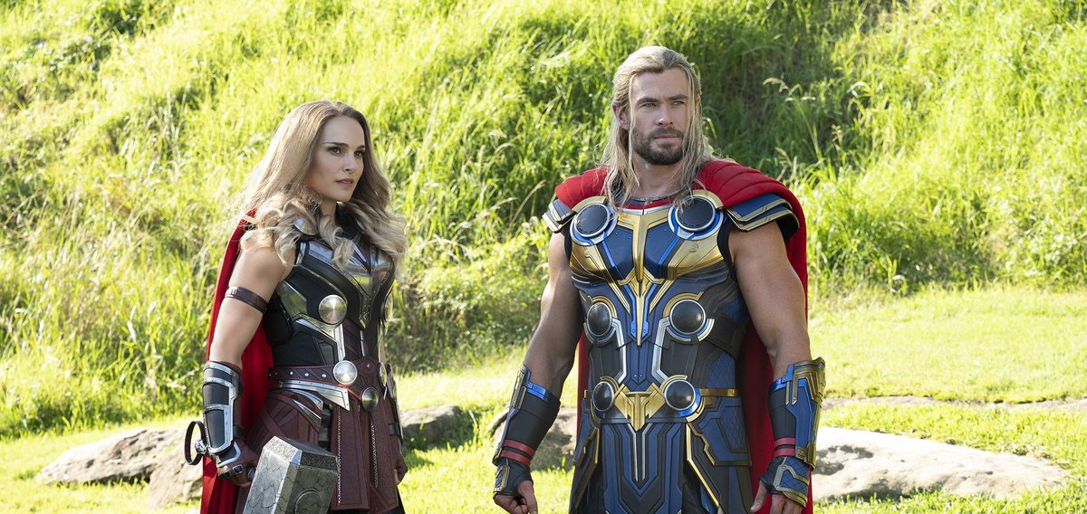 The Mighty Thor, a woman with battle armour and a red cape, along with blonde hair, holding a hammer looking into the distance, standing next to Thor, a muscled white man in blue and gold armour with a red cape