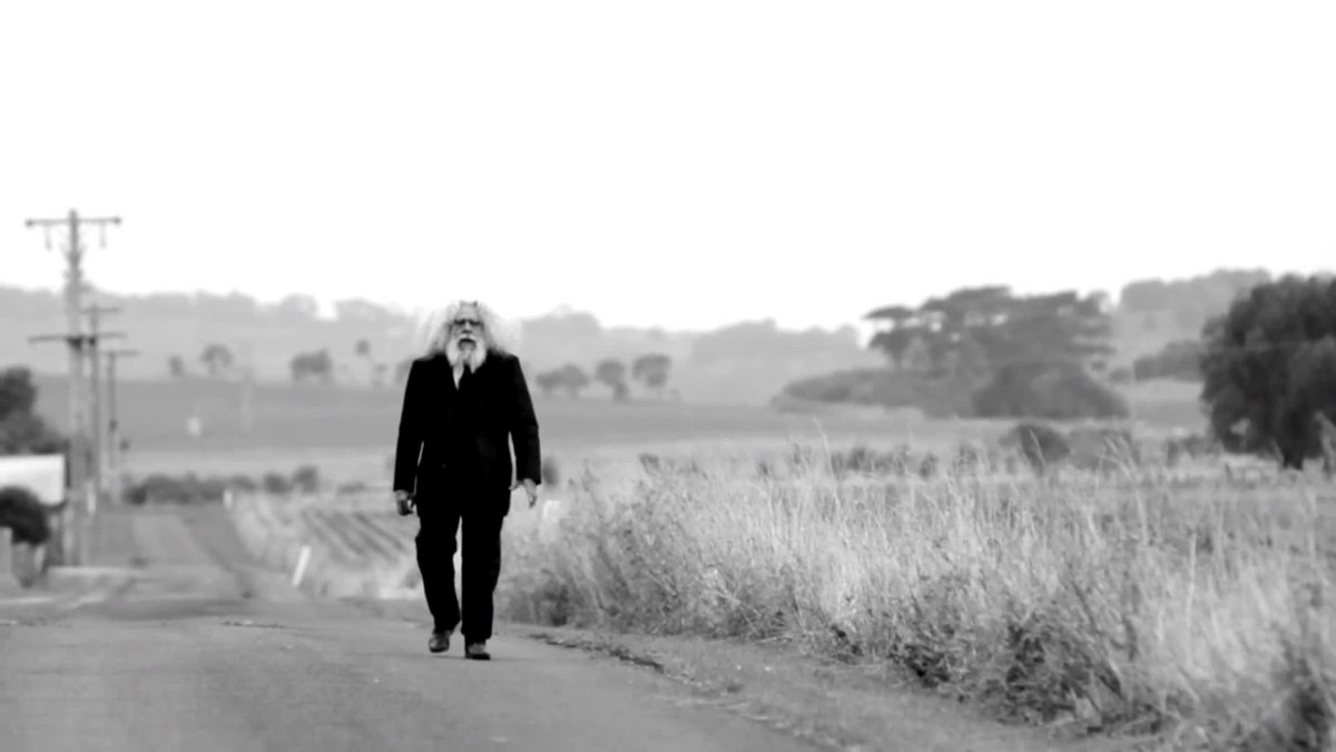 An Indigenous Elder, Uncle Jack Charles, walking down a country road in a black suit next to tall grass in black and white