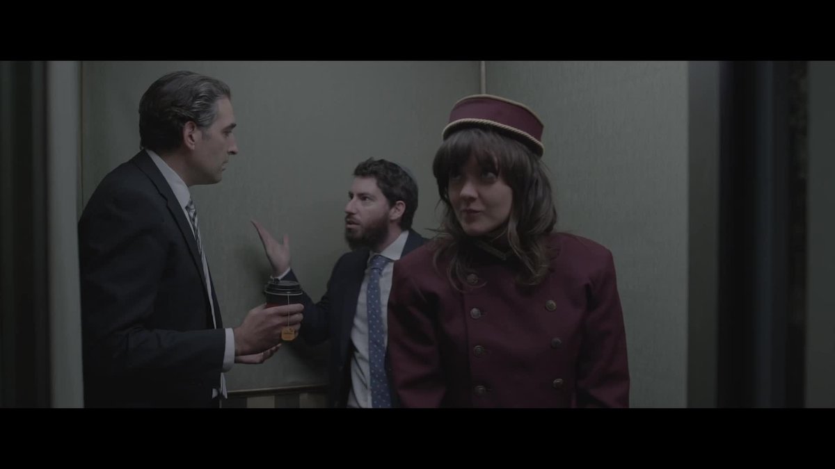 Courtney Barnett dressed as an eleavtor operator in a purple jacket and hat with two men in suits arguing behind her.