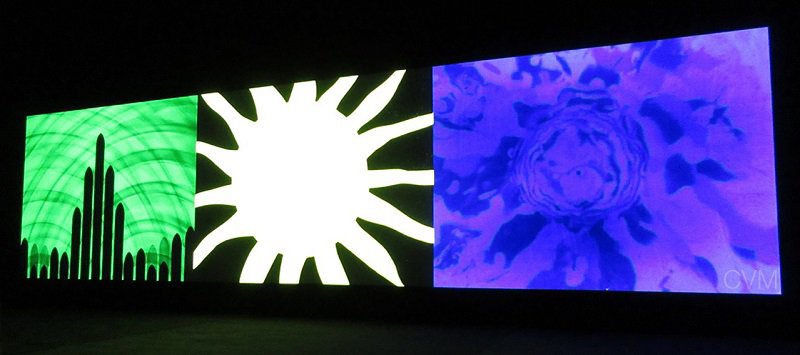 An image of a darkened room with three projected screens, each displaying different psychedelic patterns and geometric shapes