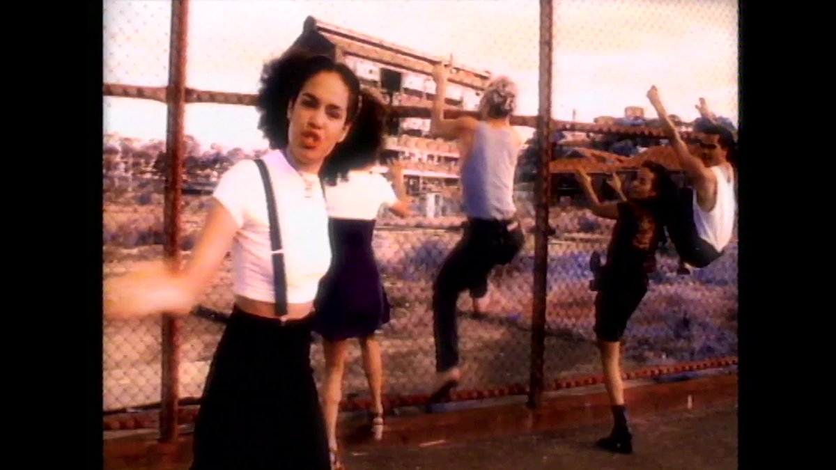 An image of Christine Anu, wearing a white top with black suspenders against a chainlink fence